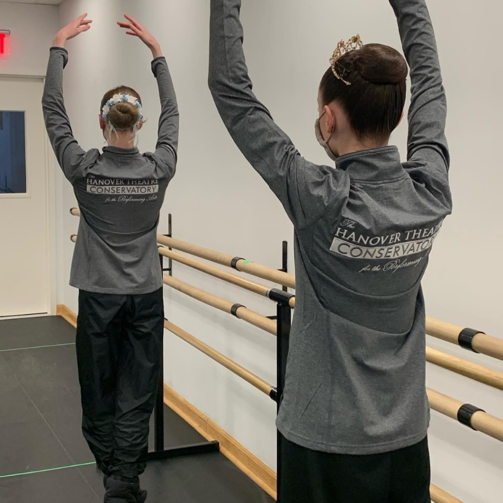 A color image of two dancers in a dance studio. They each have their hair up in a neat bun and their backs are to the camera. They are wearing warmup clothes and their jackets read Hanover Theatre Conservatory for the Performing Arts. They are in fifth position with their arms over their heads. 