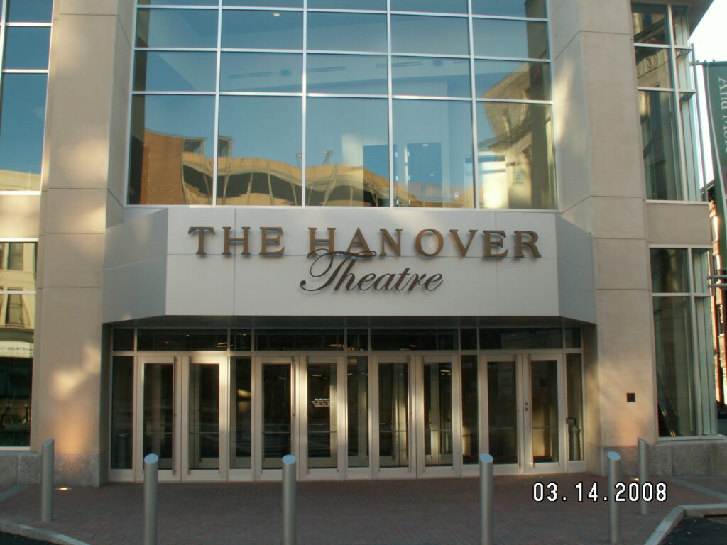 Color photograph of the entrance to The Hanover Theatre. The name is written across the front above multiple sets of doors. The section of the building we see is all windows, with reflections of other buildings in them. The date on the photo reads 03. 14. 2008.