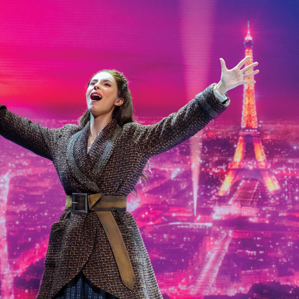 Color image of the character of Anastasia with arms outstretched, mouth open in song in front of a pink background of the city of Paris and the Eiffel Tower.