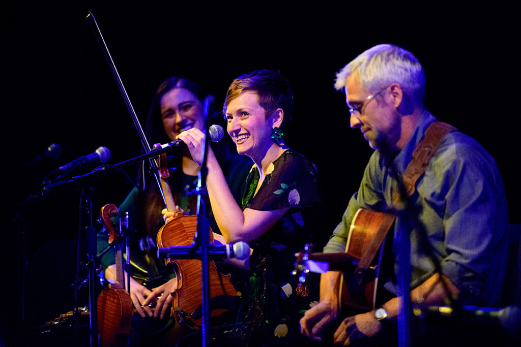 Color photo of three musicians sitting at microphones. The person on the far right is an older white man with short hair and glasses holding a guitar and looking down. The person in the middle is a white woman with short brown hair. She is holding a violin, smiling and looking out to the audience. The person on the left is a white woman with long brown hair. She is out of focus and looks smiling at the woman in the middle. 