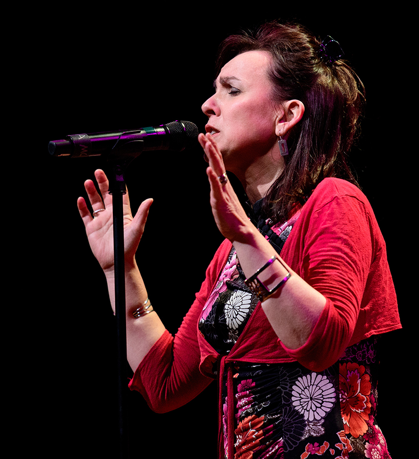 A color photo of a white woman with shoulder length brown hair that's half up. She is singing into a microphone with her hands in the air, animated. She wears a black dress with a brightly colored floral pattern and a cropped tie front red sweater. She has a big bangle bracelet on each wrist.