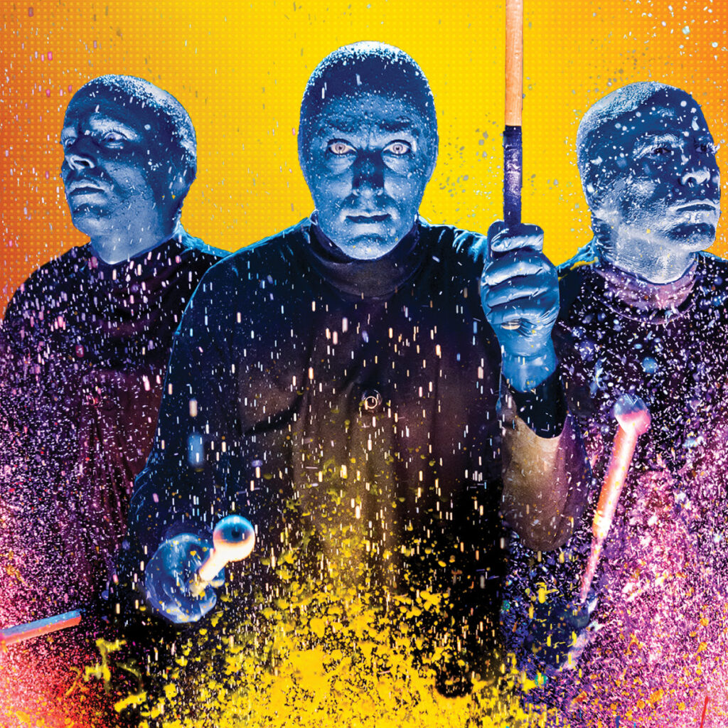Color image of Blue Man Group playing the drums as paint splashes into the air.