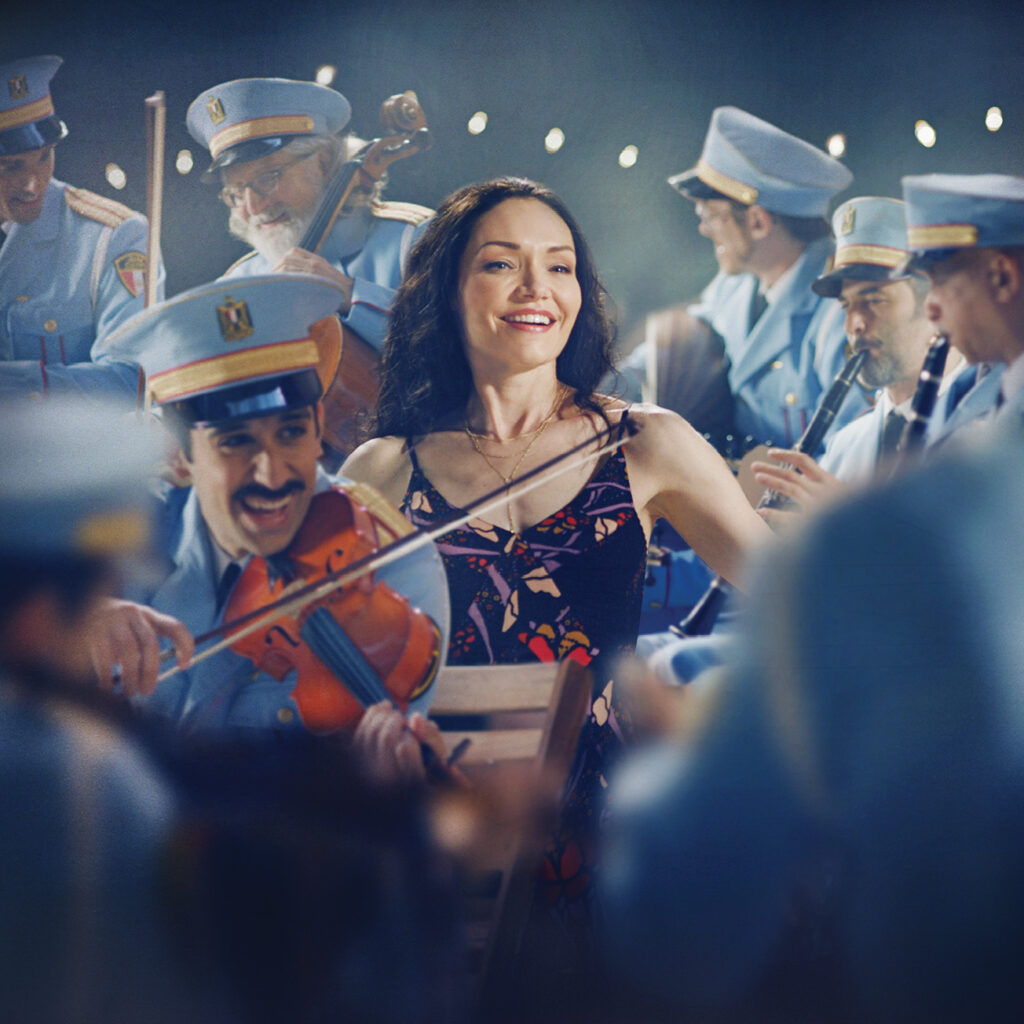 Color image of The Band's Visit featuring a female character in the center with band members in uniform playing instruments all around her. 
