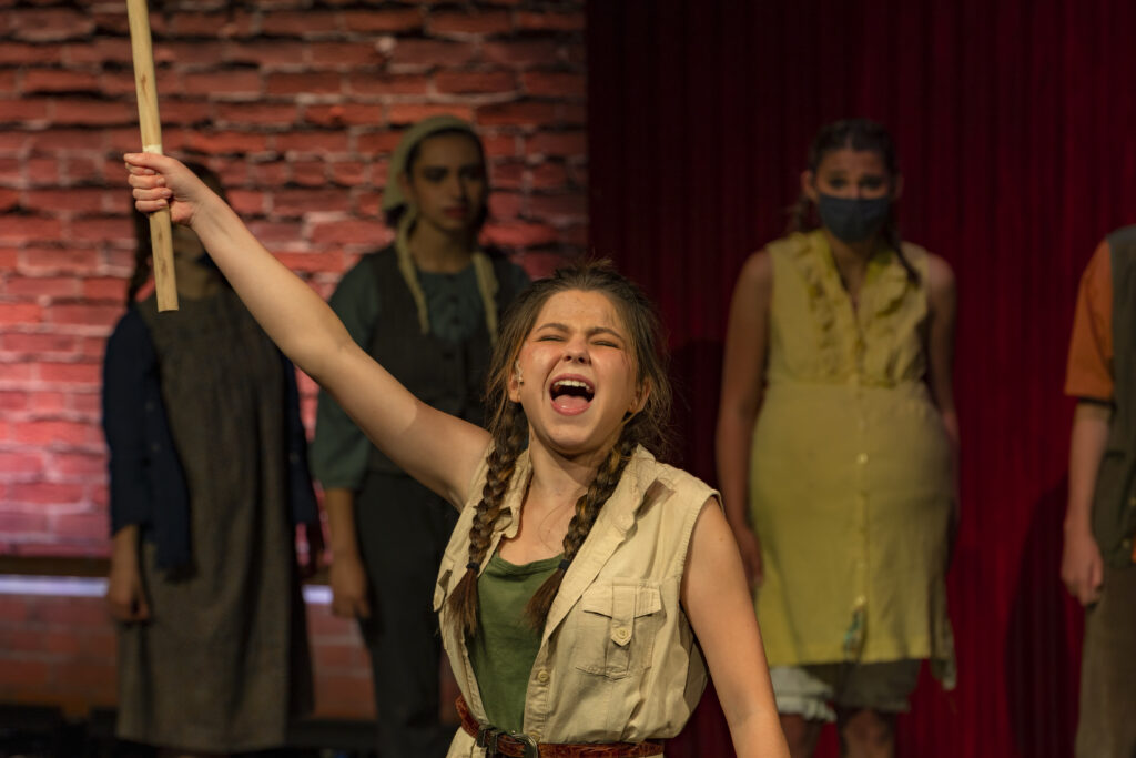 Perry Drugan, a student from the Youth Acting Company's production of Urinetown, is belting a song as she holds up a plunger. Her eyes are shut and her face is clenched as she sings. She is wearing a green shirt and a tan vest.