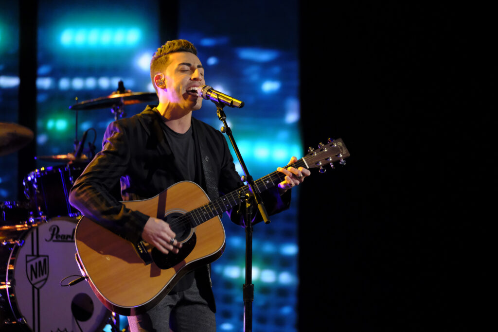 Ricky Duran is performing on "The Voice" stage in front of a blue screen and a drum set. He is wearing a black blazer, a black t-shirt and dark jeans. He is holding an acoustic guitar as he sings into a microphone. 