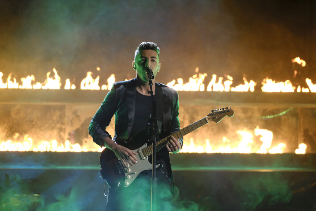 Ricky Duran is performing on "The Voice" stage in front of two lines of fire. He is wearing an all black suit  and holding a black bass guitar as he sings into a microphone. 
