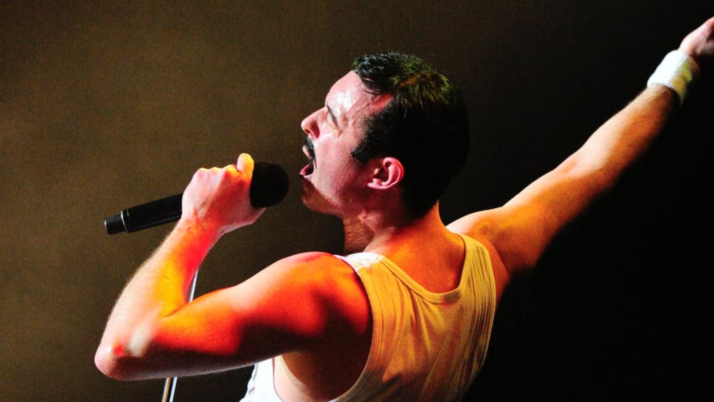 Gary Mullen is gripping a microphone, singing strongly into it. He is dressed like Freddie Mercury, with a white tank top, wrist band and signature mustache.