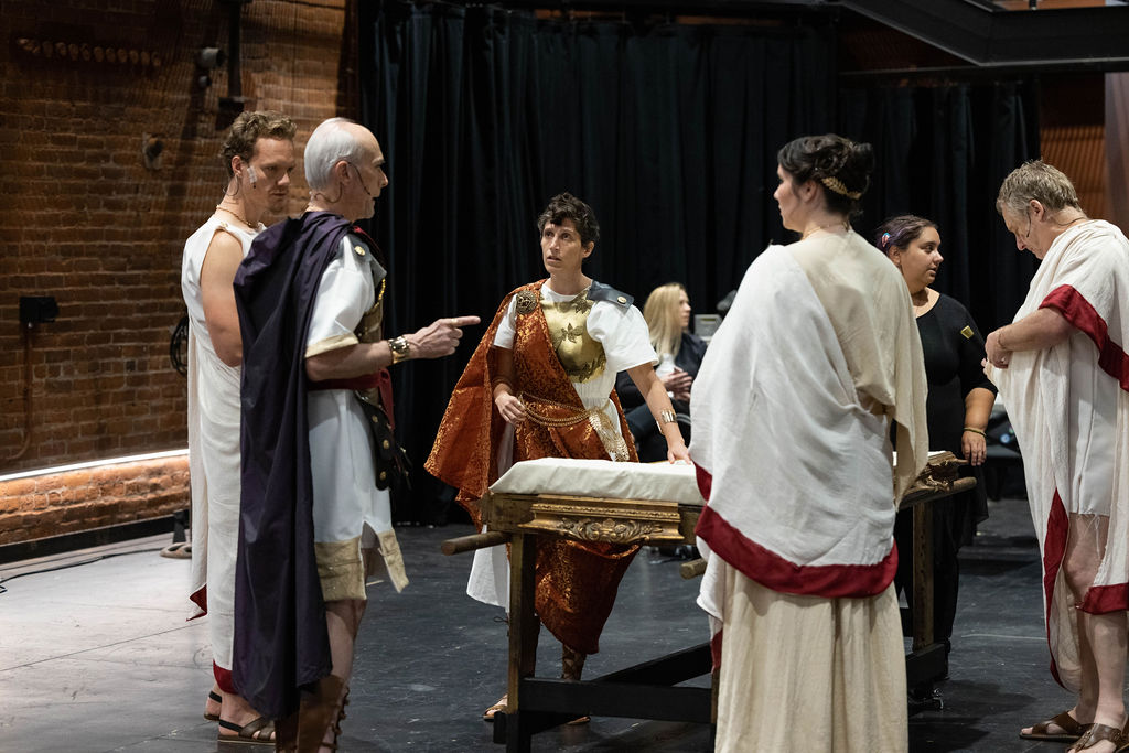A few Julius Caesar Cast members are standing in the Brick Box Theater, working on a scene. Liv Scanlon, Brandon Grimes, Dale Place, Lia Madeline and Steven Barkhimer are standing in front of the platform that Caesar's body will be carried away on in the show. They are all in full costume, in long flowing Roman garb.