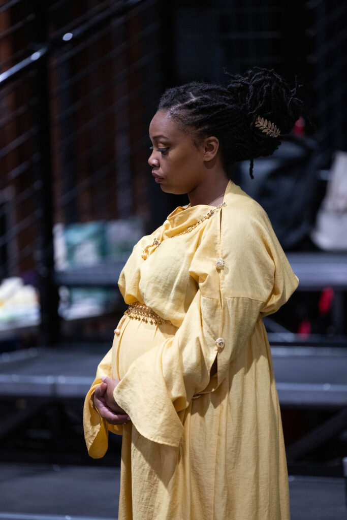 A shot of Lizzy Brooks, a black woman with elegant braids pulled up in a bun. She is wearing a yellow dress with gold accents, and she is holding her stomach as she looks to the floor in the JMAC Brick Box Theater during rehearsal.