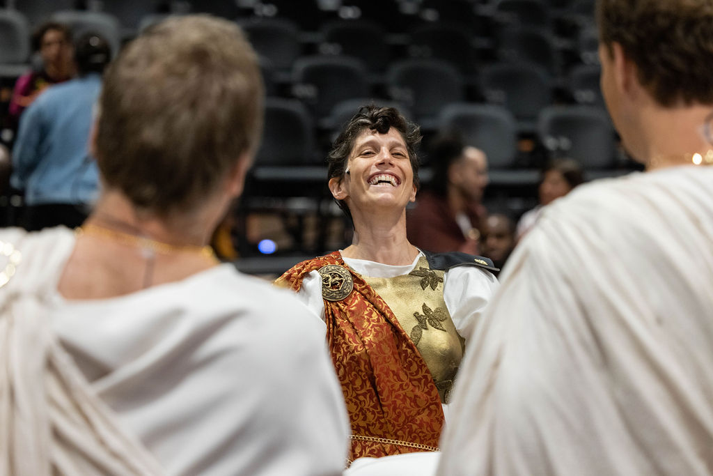 Liv Scanlon, a white woman with short curly brown hair, is standing in front of two Julius Caesar cast members. She in full costume, with a gold chest plate and a red cape. Her head is tossed back, mid laugh as she chats with actors in the JMAC Brick Box Theater.