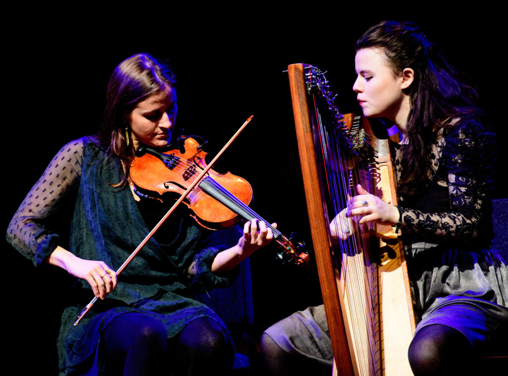 A color photograph of two women with brown shoulder length hair. The woman on the left wears a loose dark green knee length dress with sheer long sleeves and plays the violin. The woman on the left wears a black long sleeve lace top and a grey knee length skirt and black nylons. She plays a harp.