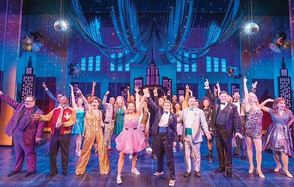 Color image of the cast of The Prom onstage, posing mid song with arms outstretched. 