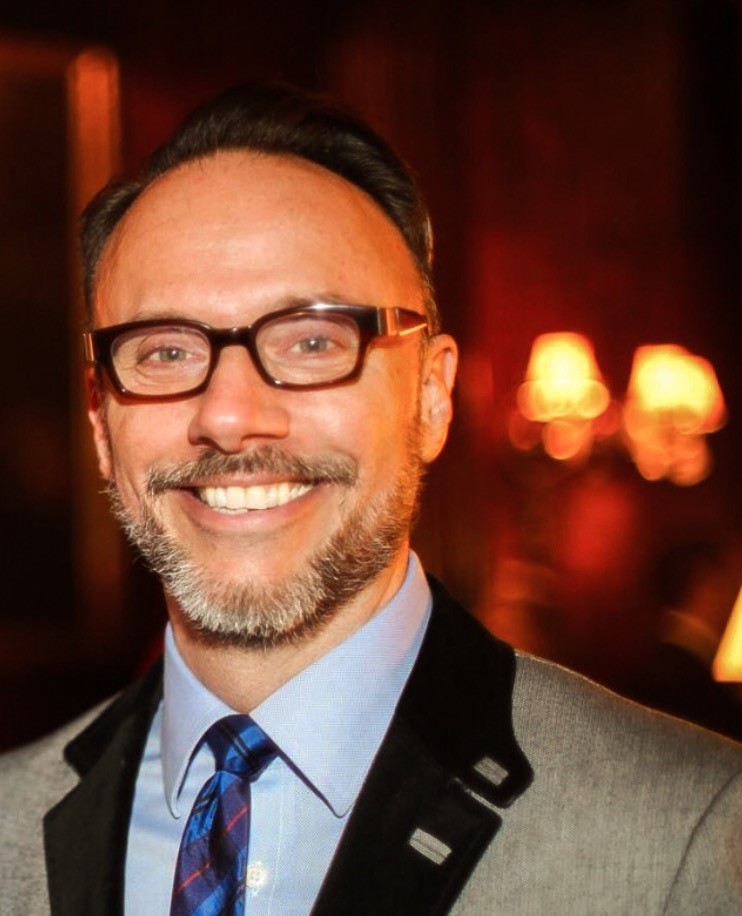 Color headshot of Chris, a white man with brown hair and facial hair. Chris is wearing a suit jacket, button-up and tie. He has glasses on and is smiling. The background is warm, out of focus tones. 