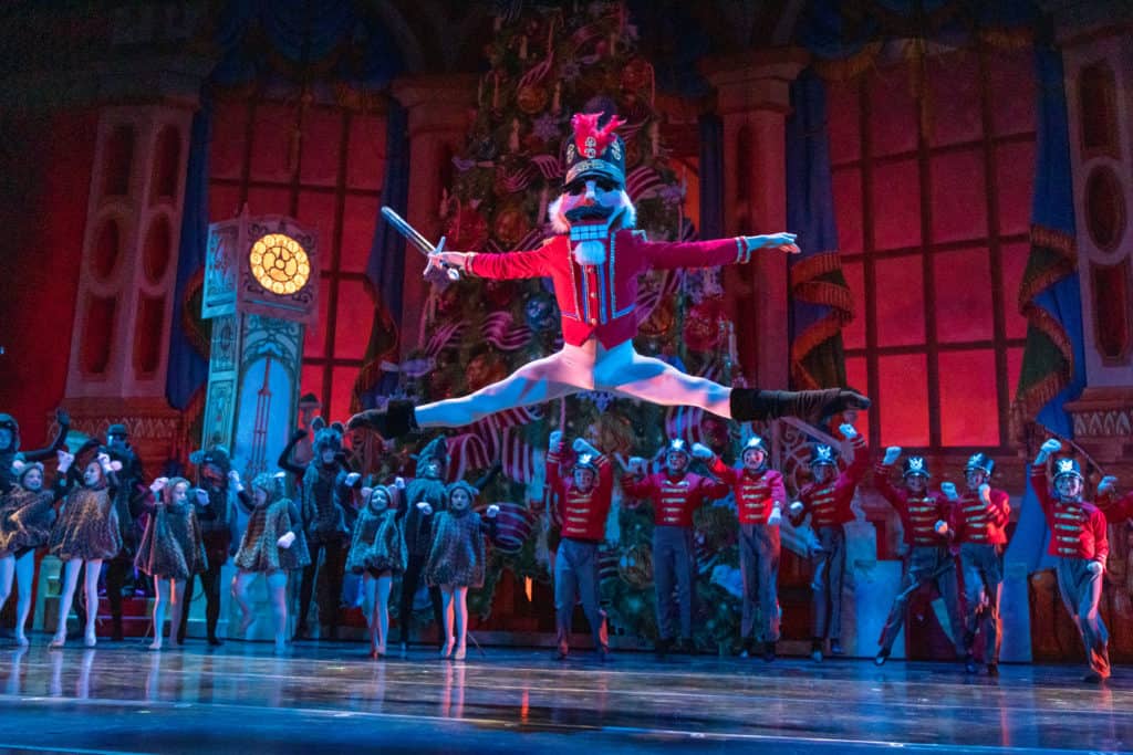 A photo from The Hanover Theatre's production of The Nutcracker. The Nutcracker is leaping across the stage with young dancers dressed as mice and nutcrackers standing behind them.