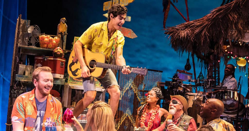 The company of the national tour of Escape to Margaritaville are surrounding a bar that Tully, played by Chris Clark, is standing on as he plays an acoustic guitar and sings.