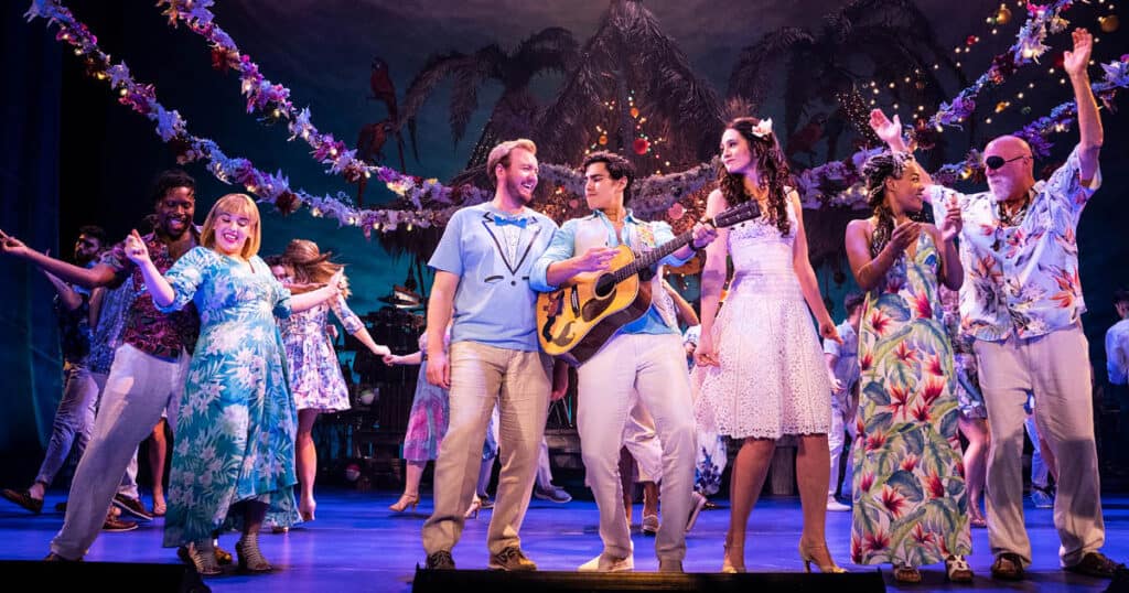 The company of the national tour of Escape to Margaritaville are dancing at the edge of a stage, wearing tropial attire as they clap, strum a guitar and throw their arms in the air.