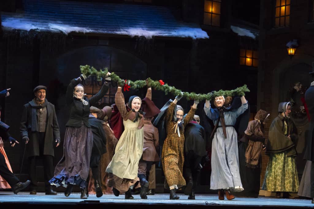 Four girls from the 2019 cast of A Christmas Carol are running downstage holding evergreen Christmas garland over their heads. 