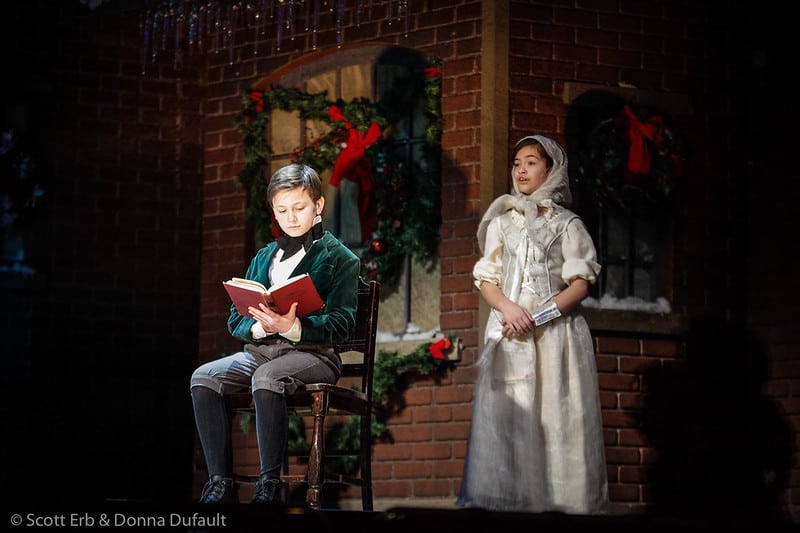 Two child actors are on performing on the stage. One of the actors is sitting in a chair and reading a book, wearing a green velvet jacket. The other actor is standing and wearing a full outfit of white.
