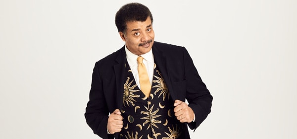 Neil deGrasse Tyson is standing in front of a white backdrop. He is holding the lapel of his jacket, showing off his space vest as he leans to the side and smiles inquisitively at the camera.