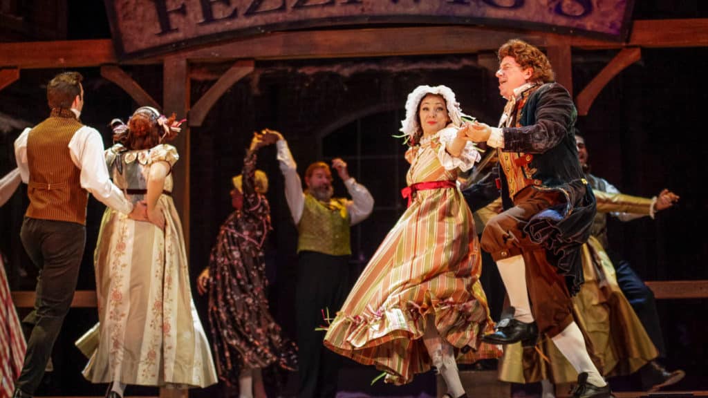 Actors on stage dressed in 1800's clothing are dancing and spinning each other in circles.