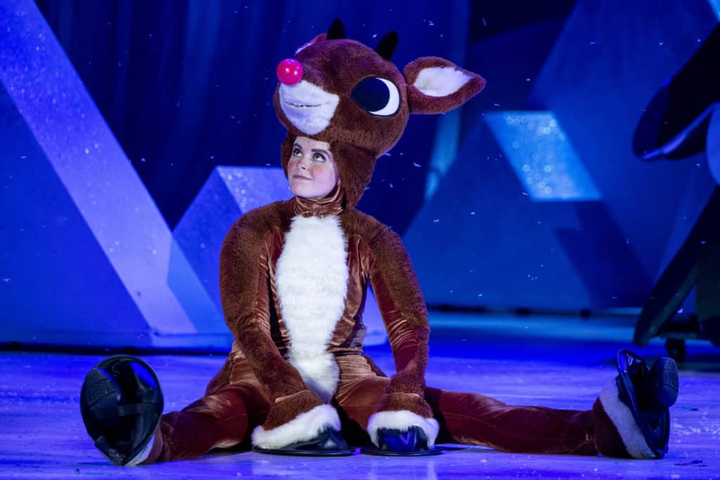 Rudolph, played by Natalie Holt MacDonald, is sitting on the floor, looking off camera at someone with a small smile.