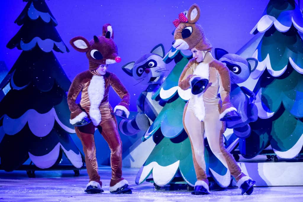 Rudolph and Clarice are standing together on a snowy stage made to look like a north pole forest. Two raccoon puppets are peeking out from behind a tree.