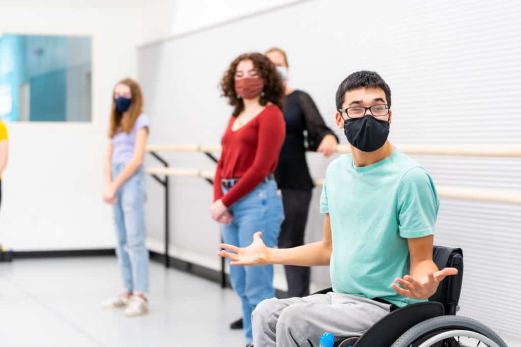 Students are participating in a summer class at The Hanover Theatre Conservatory. The students are all wearing masks. The student closest to the camera is using a wheelchair and is gesturing with his arms. The other students in the photo are standing against the wall with their hands clasped. 