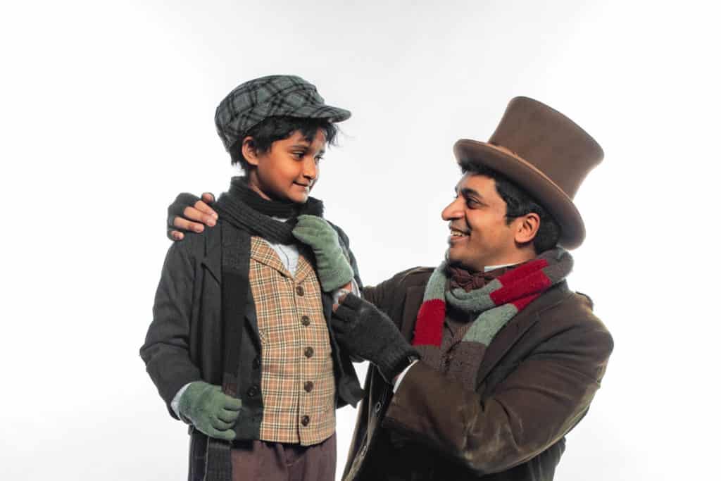 Sri Chaitra Prasadula and Sriram Emani are posing for a picture dressed as their characters, Tiny Tim and Bob Cratchit respectively. Sri is wearing a plaid shirt, a plaid vest and winter accessories. Sriram is wearing a top hat, finger-less gloves, and other winter accessories. Sriram has his arms around Sri in a fatherly pose.