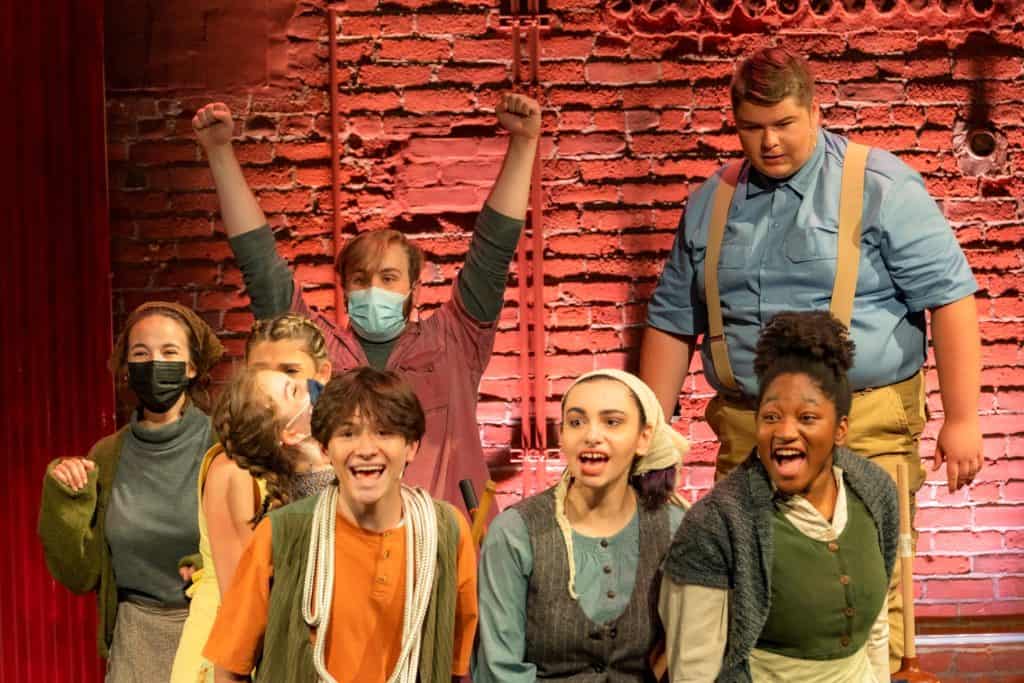 Eight performers are acting in Urinetown. Some performers are wearing masks and others have their mouths open to sing. They are wearing neutral and Earth-tone colors.