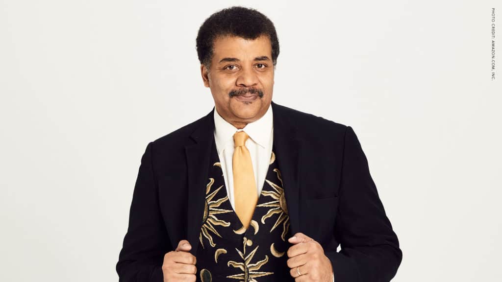 Neil deGrasse Tyson is standing in front of a white backdrop. He is holding the lapel of his jacket, showing off his space vest as he smiles at the camera. 