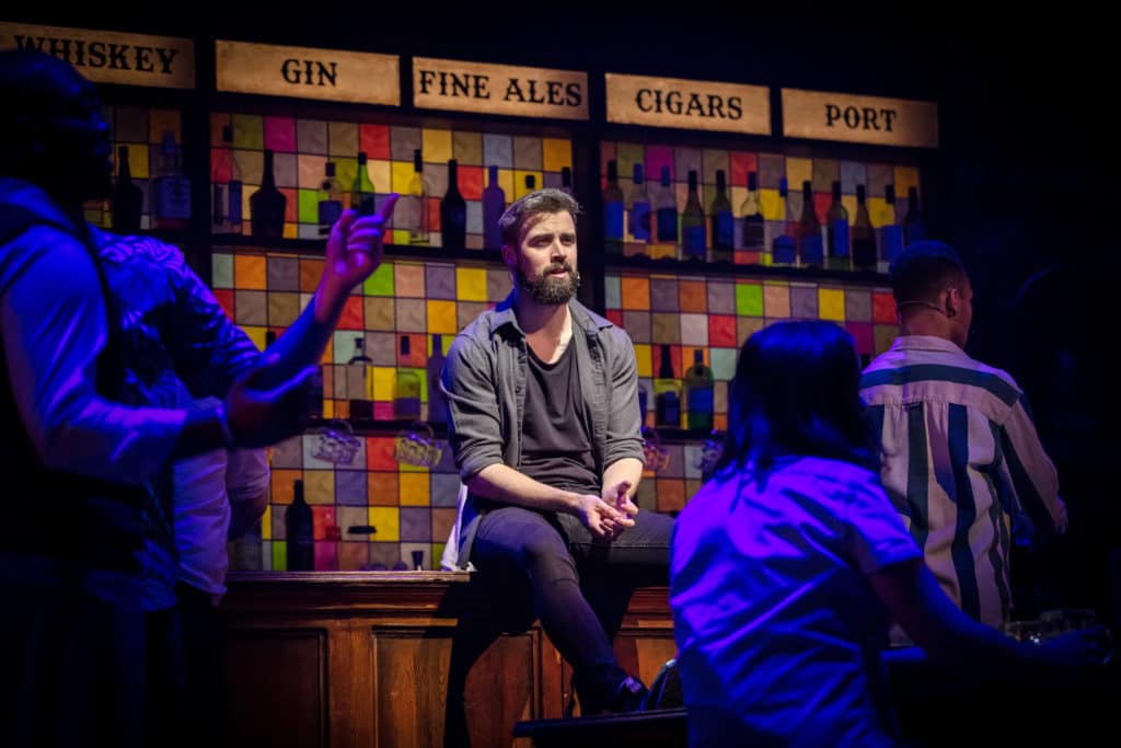 Mark Irwin, wearing a grey button down shirt, is sitting on the pub set. He is surrounded by bottles and colorful tiles, and the spotlight is shining on him. 
