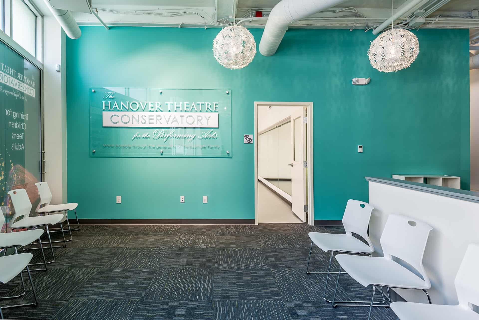 A glass sign that reads "The Hanover Theatre Conservatory for the Performing Arts" hanging on a blue wall, a studio can be seen in the background, chairs line the wall on the right side of the room