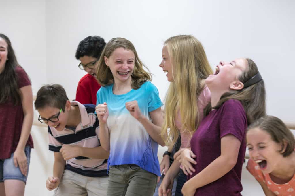 A group of students in Acting class laugh and smile. The students are standing in a line in a white room, and some are in-motion, as if they are dancing. 
