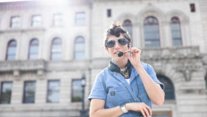 Liv Scanlon stands in front of the Worcester City Hall wearing sunglasses and a microphone