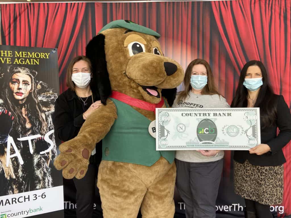 Country Bank's mascot Buck, a big brown dog with a green vest and cap, is standing alongside Lisa Condit, Kristin Mullins and Jodie Gerulaitis. Kristin and Jodie are holding a Country Bank $1,000 bill photo prop. 