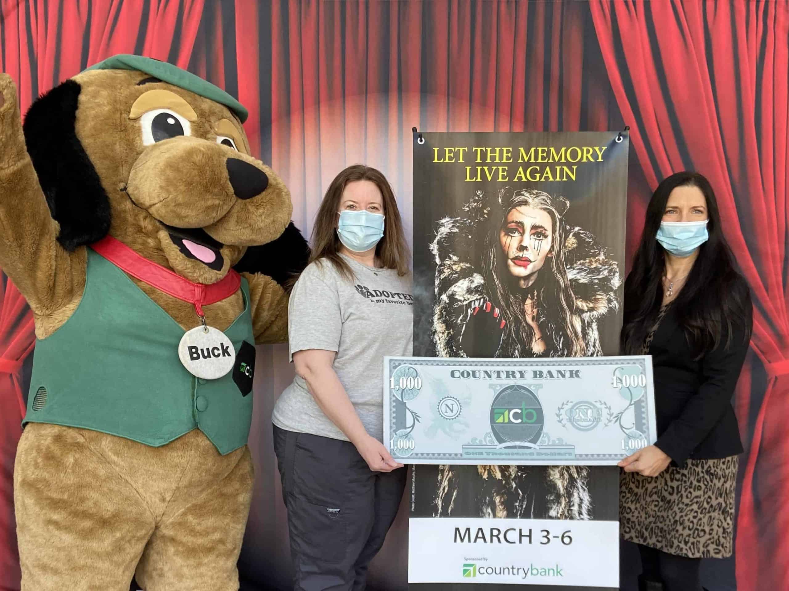 Country Bank's mascot Buck, a big brown dog with a green vest and cap, is standing alongside Kristin Mullins and Jodie Gerulaitis. Kristin and Jodie are holding a Country Bank $1,000 bill photo prop. 