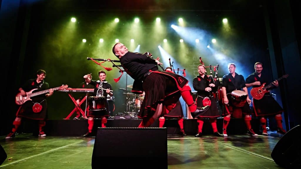 A member of The Red Hot Chilli Pipers is performing with his bag pipes marching across the front of stage. The band ensemble is wearing black polo's with red plaid kilt's and they're lined up right behind the main performer.