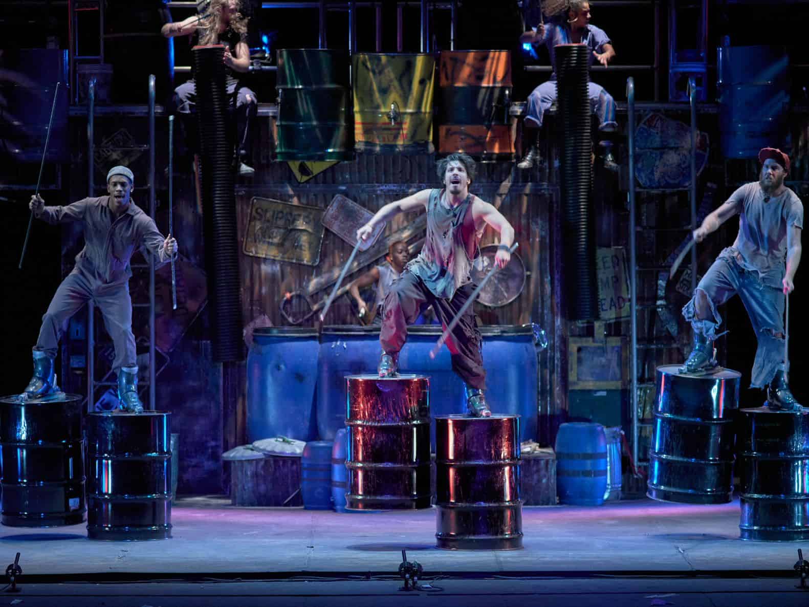 Performers from STOMP appear on an open industrial stage. Three performers are downstage standing on top of aluminum trash cans with large drum sticks in the midst of a dance. They are wearing clothing in a gray color scale with a distressed aesthetic.