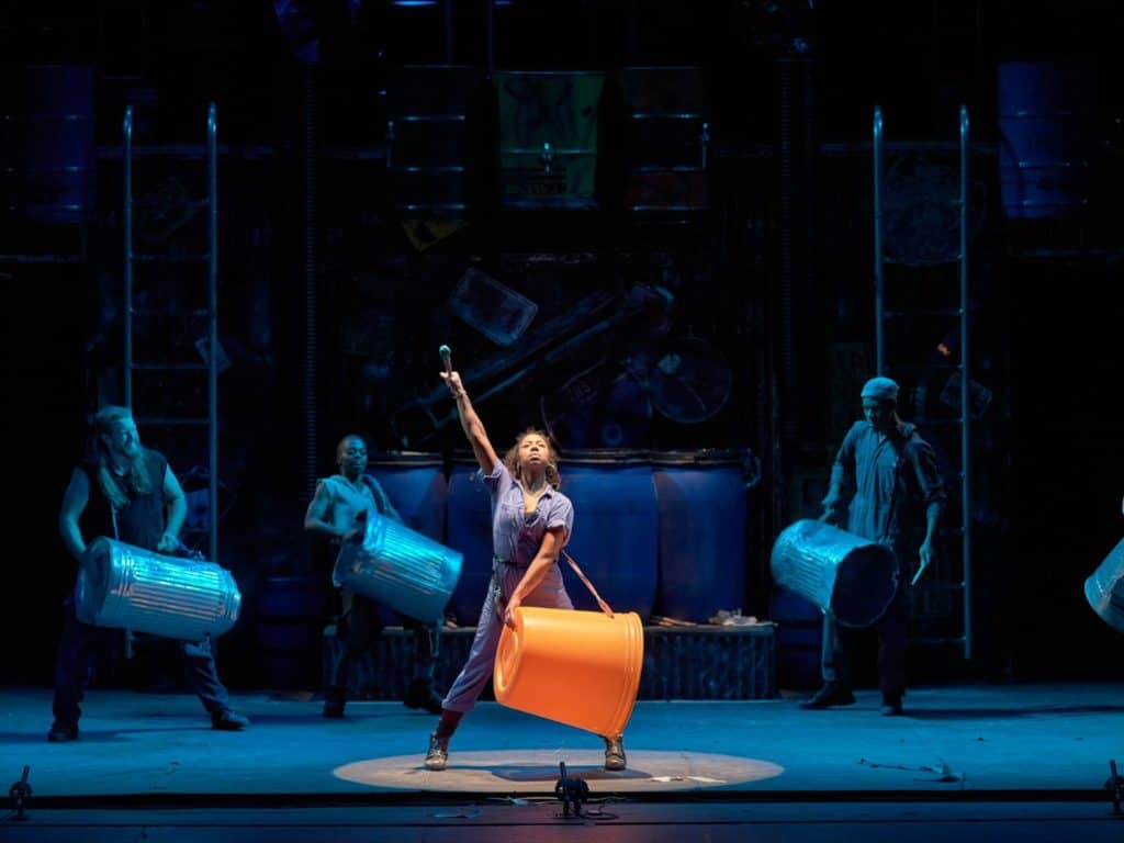 A female performer is standing underneath a spotlight wearing a blue utility jumpsuit with a defiant facial expression. She is carrying a large orange bin and has a drum stick up in the air. Behind are other members of the compony carrying trash cans and drum sticks as well, standing in a semi-circle.