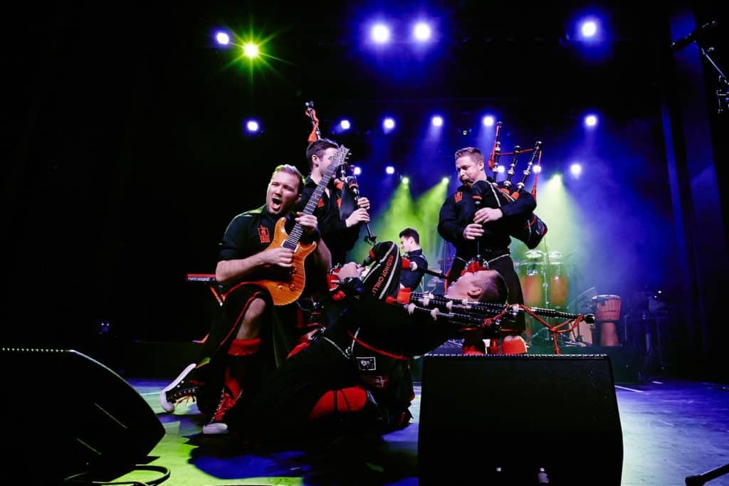 The Red Hot Chilli Pipers are in the midst of one of their bag-rock performances. Out of a group of four performers on the main stage, three members are playing bag pipes while the other is playing electric guitar.