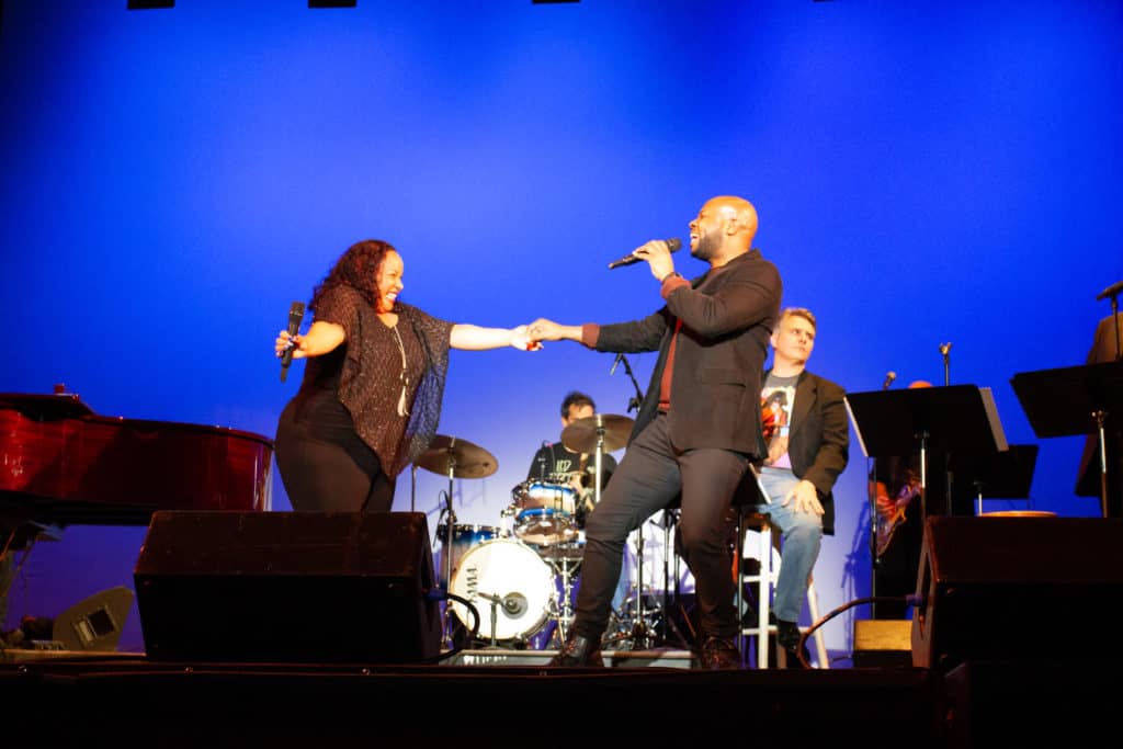 Two performers are holding hands as they sing to one another in front of the 50 Years of Rock and Roll band.