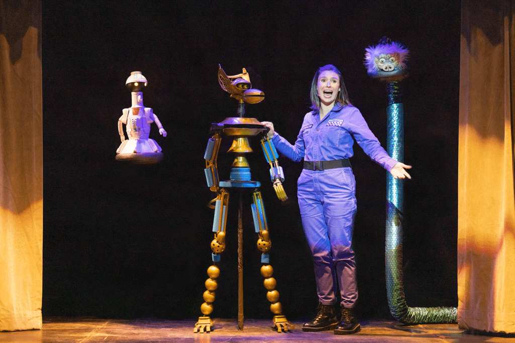 Emily Connor is standing alongside the MST3K robots Tom Servo, Crow T. Robot and GPC. Her arm is outstreched as she talks to the crowd.