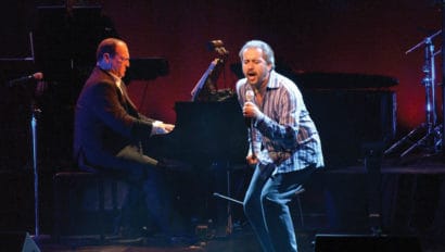 Two men are performing on stage. One man is performing with a microphone and is singing. The other man is sitting at the piano.