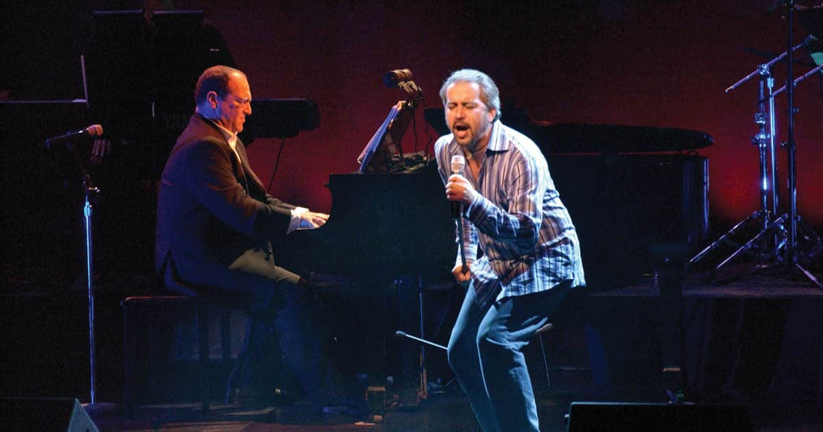 Two men are performing on stage. One man is performing with a microphone and is singing. The other man is sitting at the piano.