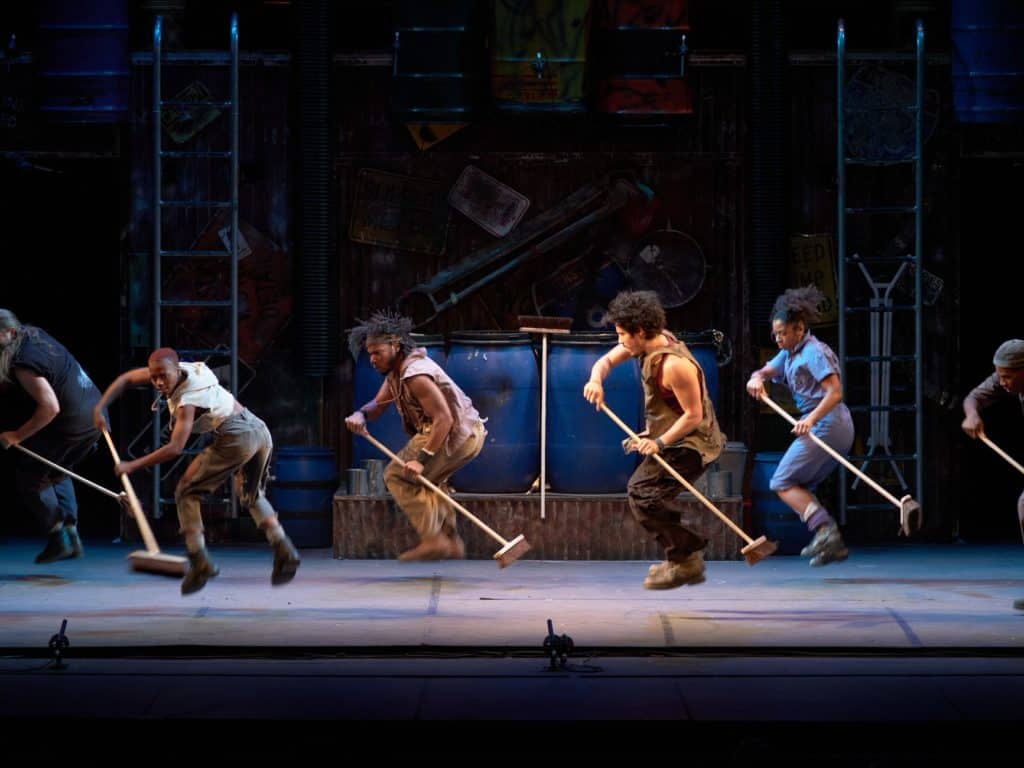 Six performers of the cast are in the midst of a dance number doing a jump in the air while holding a broom.