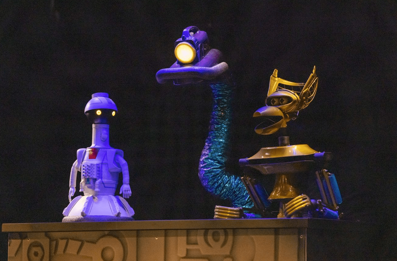 Three robots sit behind a counter. Tom Servo, Crow T. Robot and GPC sit together on stage. Tom Servo is standing on top of the table.