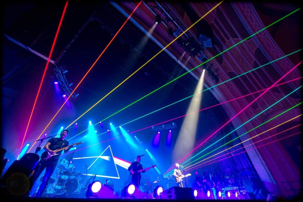 Members of Brit Floyd are in the midst of a musical performance with the prism of light "Dark Side of the Moon" artwork displayed on a screen. Various laser beams in vivid primary colors are casted in the audience.