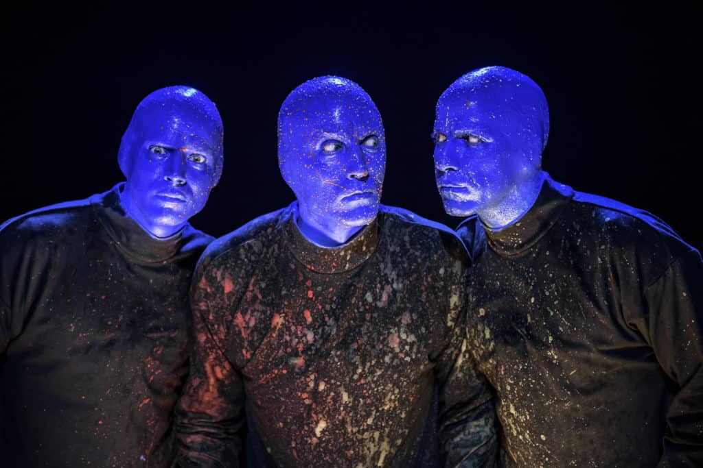 Three Blue Men from the Blue Man Group stand in a row. The men have blue skin and colorful paint splattered on their faces. They are wearing the same black shirts that are also splattered with paint.