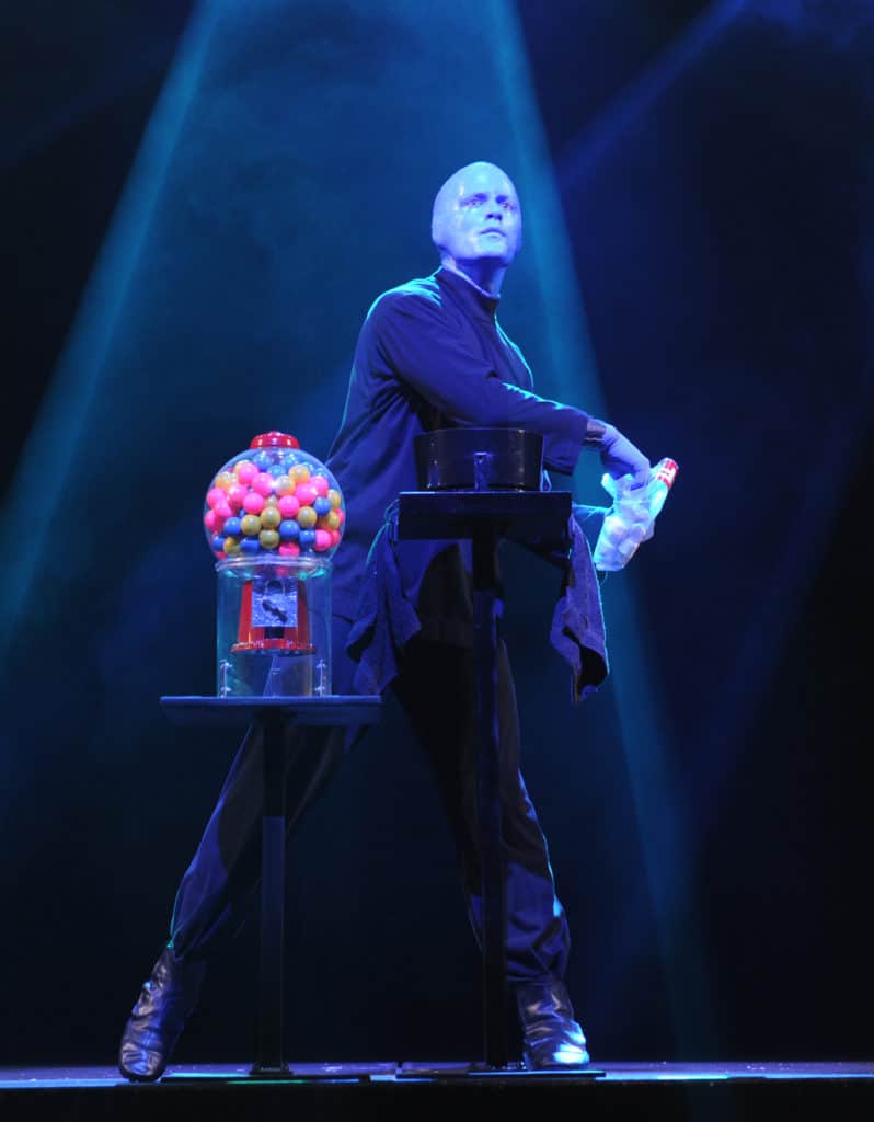 One of the Blue Man does the "toss and catch" with the audience. He is standing on stage with a blue light cast over him. The blue man is standing next to a gumball machine, and he is throwing marshmallows out into the audience.