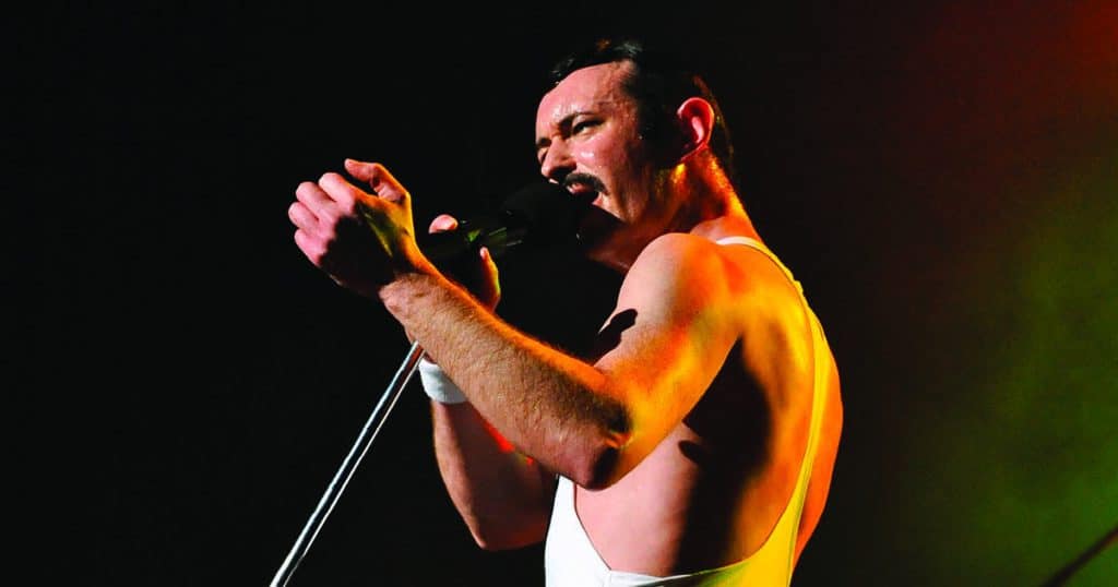 Gary Mullen is gripping a microphone, singing into it as he looks off in the distance. He is dressed like Freddie Mercury, with a white tank top, wrist band and signature mustache.